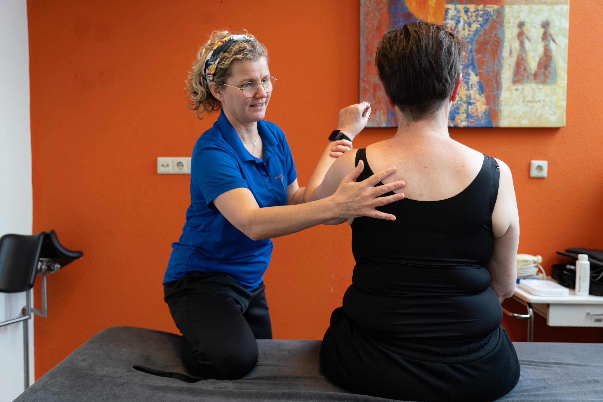 Oedeemfysiotherapie. Patiënt zit met een uitgestrekte arm op het behandelbed, terwijl de fysiotherapeut haar arm observeert.