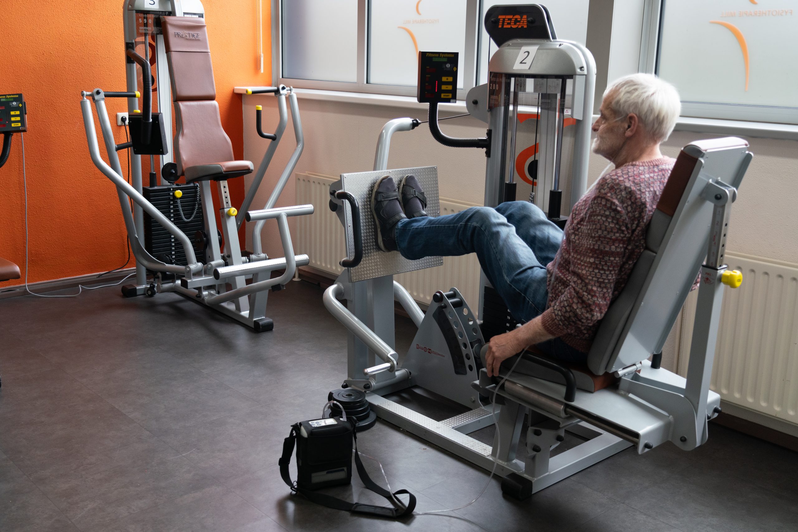 Man met zuurstofbox, die op fitnessapparatuur zit voor longfysiotherapie.