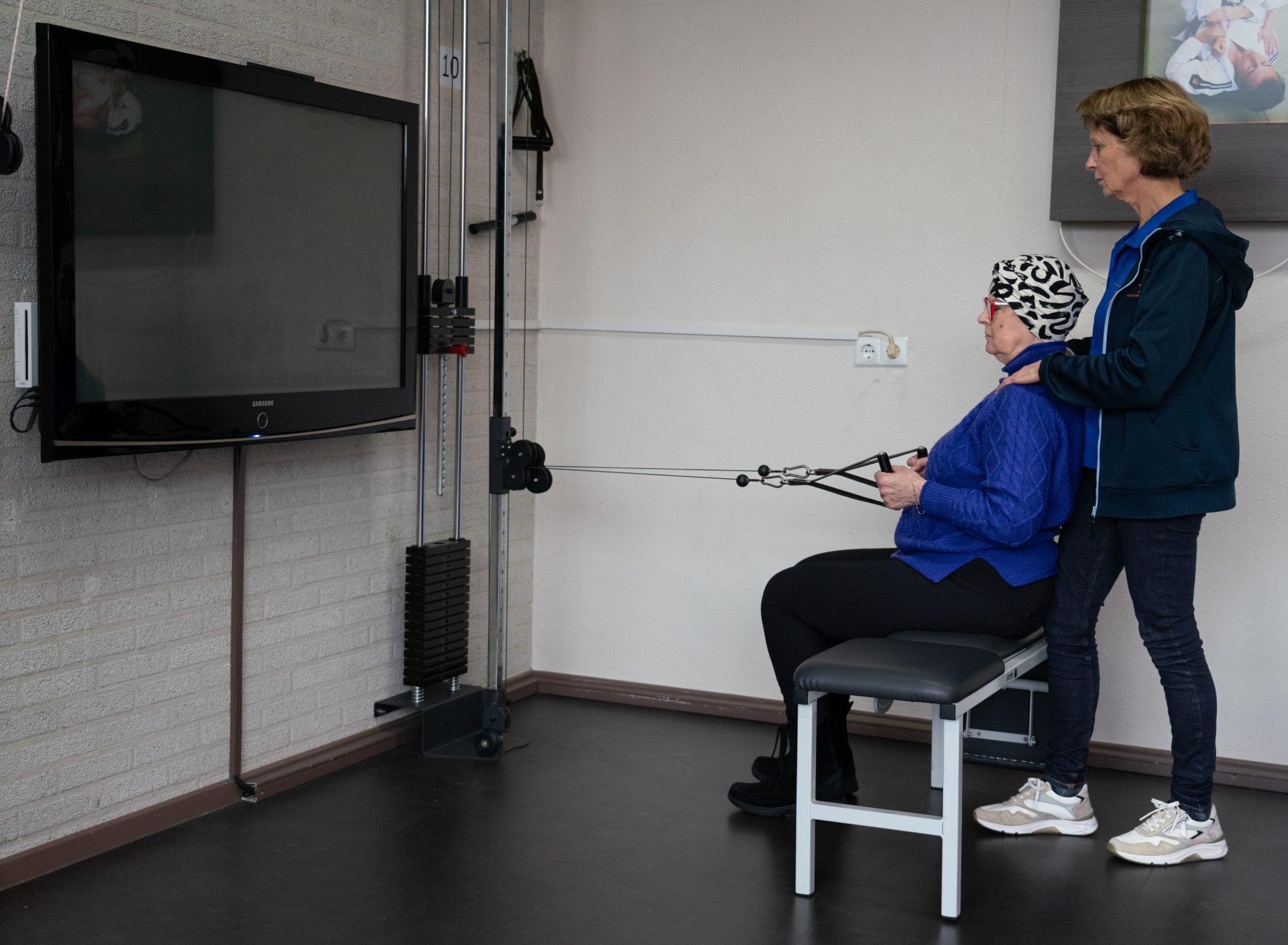 Oncologiefysiotherapie. Een vrouw is kabelopdrachten aan het doen, terwijl de fysiotherapeut haar begeleidt.